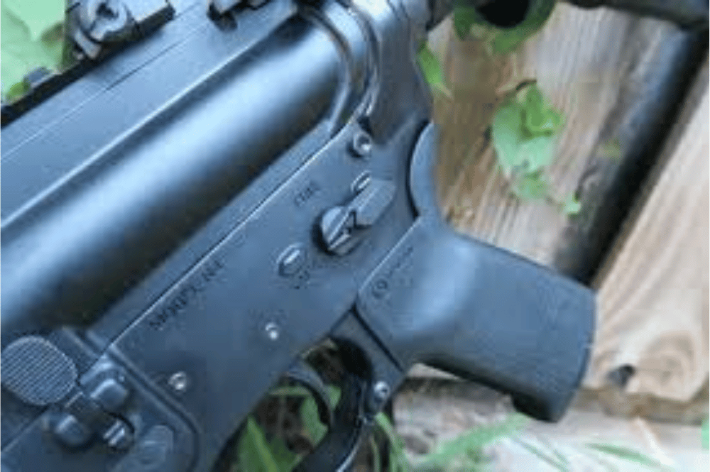 An ambi safety in an AR15 shown sitting against a fence on tile with some vines
