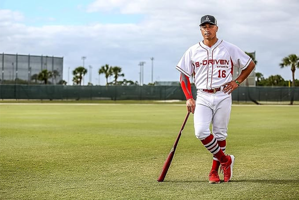 A baseball player leaning on his bat in the outfield wearing a red arm sleeve.
