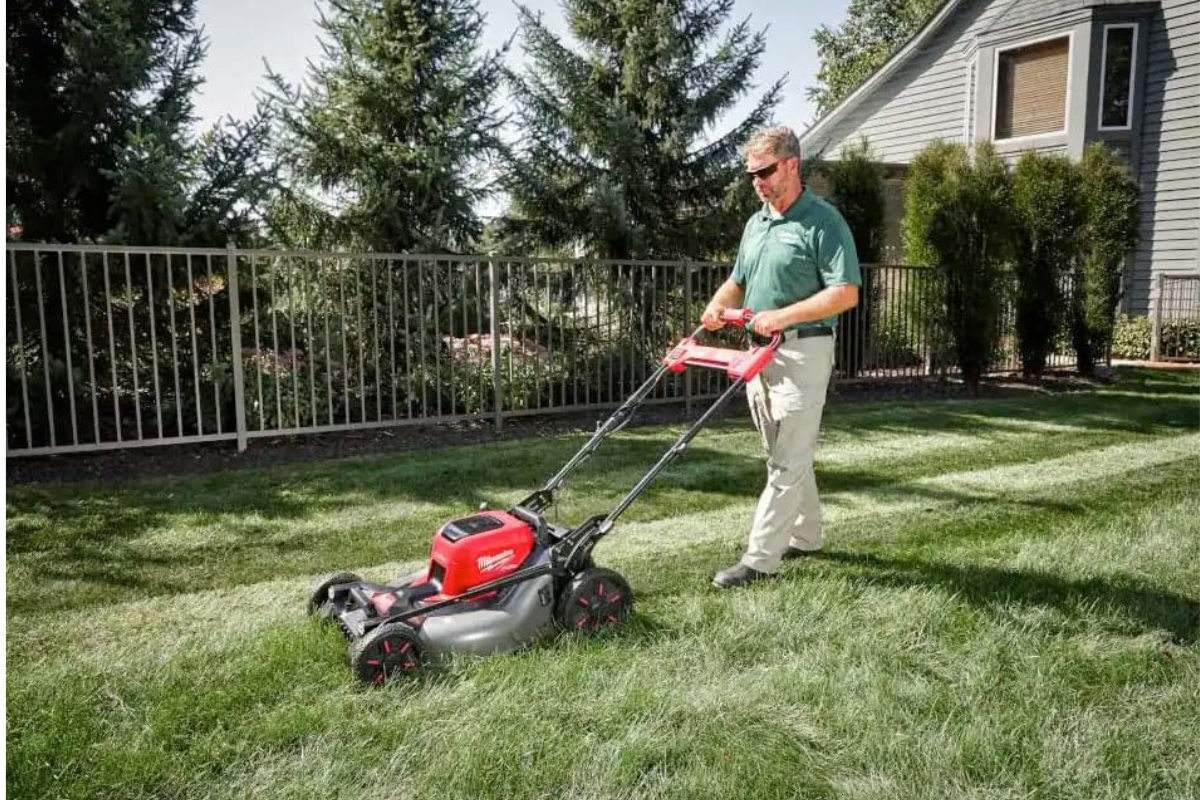 A man using a Milwaukee cordless lawn mower in tall grass in a fenced yard.