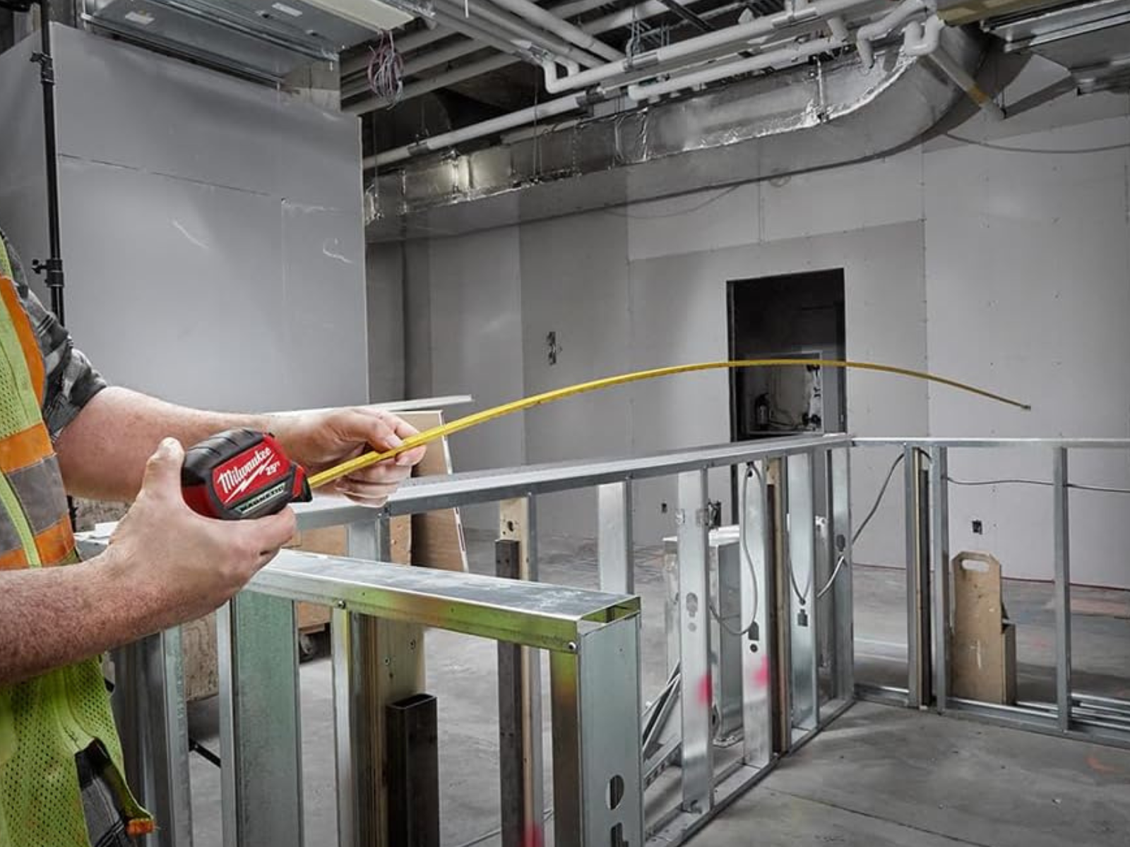 A man with a Milwaukee measuring tape extended 5 meters while building a steel frame wall inside a large building