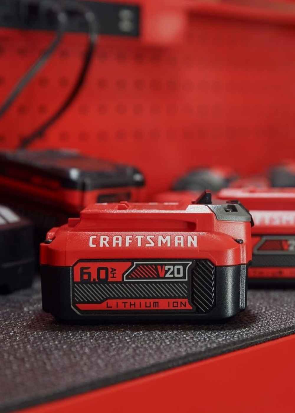 A 6.0 amp 20 volt battery by Craftsman sitting on a tool box with Craftsman chargers and batteries behind it